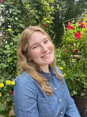 A smiling person with wavy, light brown hair wearing a denim button-up shirt sits outdoors in front of lush greenery and colorful flowers, including red and yellow blooms.