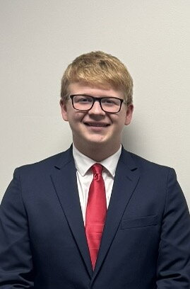 A smiling person with short blond hair and glasses wearing a navy blue suit, white shirt, and red tie stands against a plain light-colored background