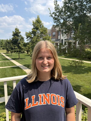 A smiling person with shoulder-length blonde hair wearing a navy blue 'ILLINOIS' T-shirt stands outside on a sunny day, with green lawns, trees, and brick houses in the background