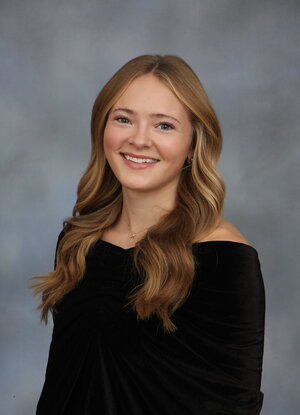 A smiling person with long, wavy light brown hair wearing a black off-the-shoulder top poses for a professional portrait against a gray studio background.