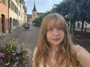 A person with long blonde hair takes a selfie on a charming street lined with colorful buildings, flowers, and trees.