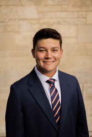 A smiling person with short dark hair wearing a navy blue suit, light blue dress shirt, and navy-and-orange striped tie poses for a professional portrait.