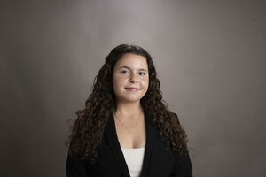 A person with long brunette hair, wearing a black blazer with a light brown background. 